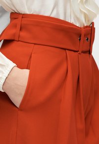 Κόκκινο, ψηλόμεσο παντελόνι από υφή υφάσματος με φαρδύ ζωνάρι και πιέτες. Διαθέτει πλαϊνές τσέπες και κούμπωμα με κουμπί.