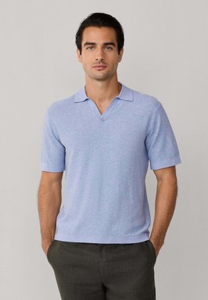 Hombre de pie con las manos en los bolsillos, vestido con una camisa tipo polo de punto azul claro de manga corta y pantalones oscuros, sobre un fondo liso.