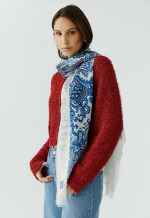 Femme portant un pull rouge texturé, une écharpe à motif paisley bleu et un jean bleu clair, debout devant un fond blanc uni.