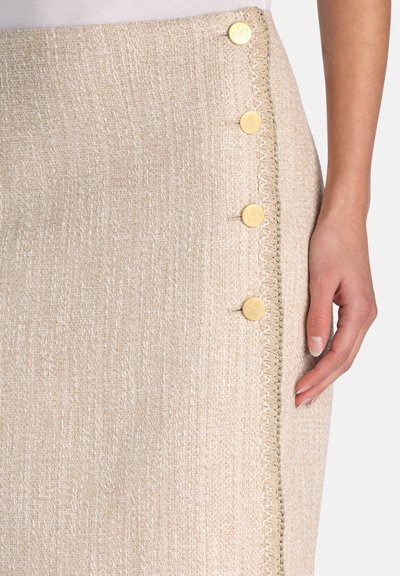Beige textured skirt with four gold decorative buttons and beige trim, shown with a hand resting beside it.