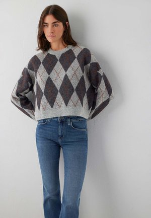 Frau trägt einen grauen Pullover mit Rautenmuster und blaue Jeans, steht vor einer hellgrauen Wand mit den Händen hinter dem Rücken.