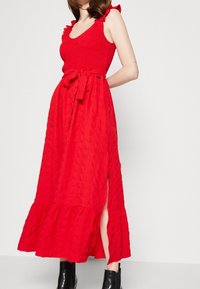 Punainen maksimekko tekstuuripinnalla, smokkikiristetyllä yläosalla, röyhelöstrapeilla ja vyötärönauhoilla. Sisältää sivupurjeen ja kerroksellisen helman. Mustat nilkkakengät.