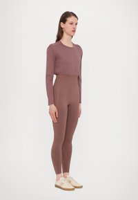 Mauve long-sleeve top paired with high-waisted mauve leggings; both in smooth fabric. Model wears light shoes with a textured sole.