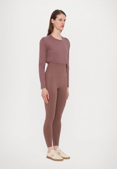 Lavendelfarbenes Langarmoberteil, kombiniert mit hochtaillierten lavendelfarbenen Leggings, beide aus glattem Stoff. Das Model trägt helle Schuhe mit einer strukturierten Sohle.