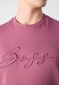 Violet trumpas rankovėmis marškinėliai su apvalia kaklo iškirpte ir išsiuvinėtu tamsesnio violetinio atspalvio "Boss" logotipu priekyje, pagaminti iš smagaus medvilnės audinio.