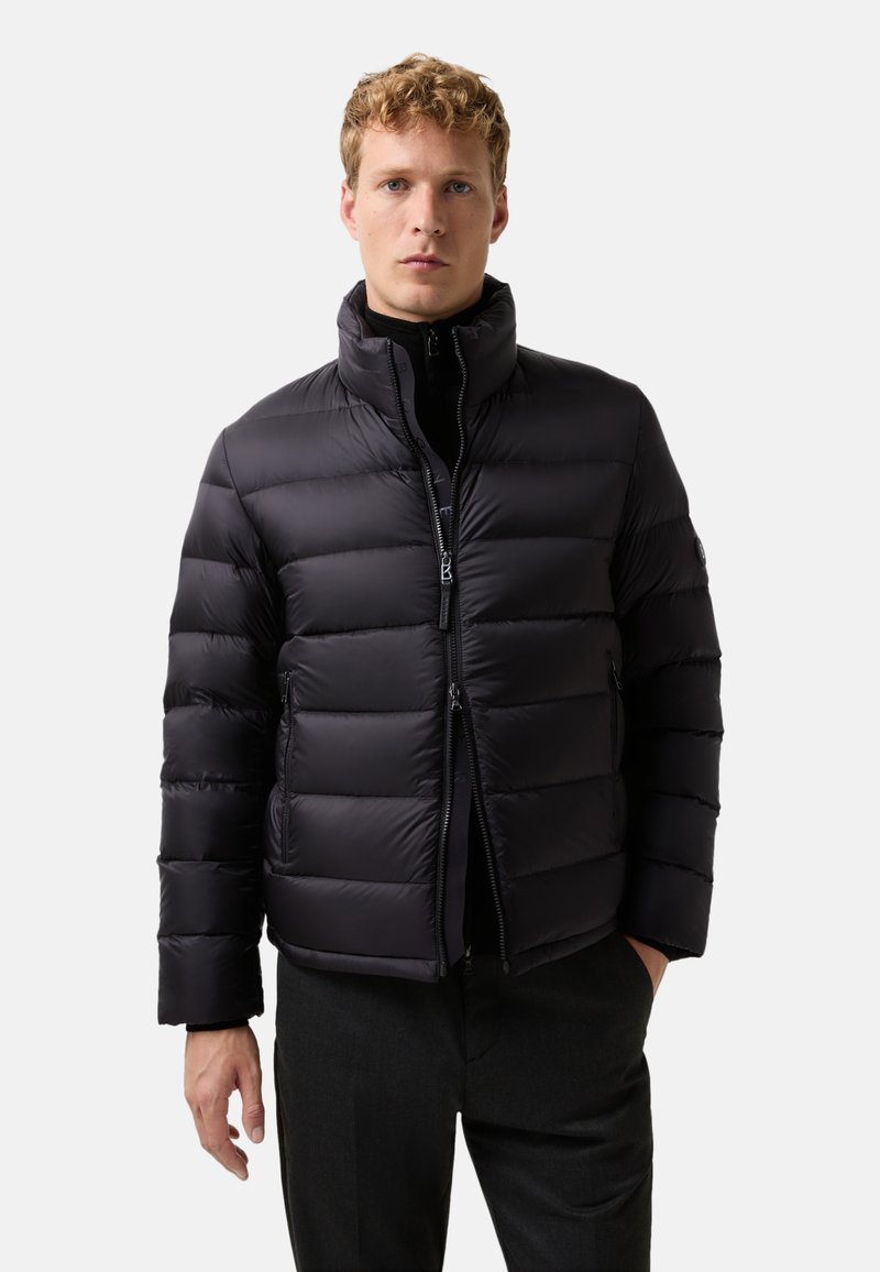 Ung mand iført en sort polstret jakke over en sort lynlåsbluse, stående med en hånd i lommen mod en ensfarvet lys baggrund.