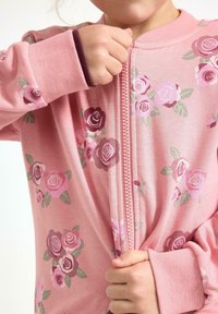 Dziecko w różowej kurtce w kwiaty, ręce zaciągające zamek błyskawiczny z przodu, zbliżenie na tułów i ramiona.