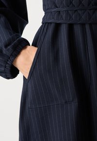 Robe bleu marine en tissu texturé, avec de subtils rayures verticales, une ceinture à nouer amovible et une poche latérale. Manches à poignets élastiques.