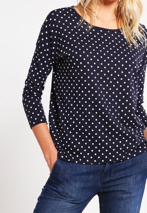 Femme portant un haut à manches longues bleu marine à pois blancs et un jean bleu, les mains dans les poches, sur un fond blanc.