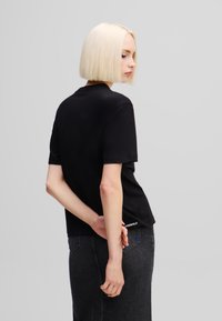 Melns īsām piedurkn�ēm t-krekls no mīksta kokvilnas maisījuma, ar apaļu izgriezumu un smalku logotipa detaļu uz apakšmalas. Modelis valkā tumšas džinsa svārkus.