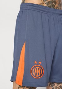 Shorts de sport bleus avec des accents latéraux orange et un logo brodé avec deux étoiles. Fabriqués en tissu texturé avec une taille élastique.