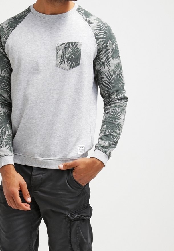 Uomo che indossa un felpa grigia chiara con maniche e tasca a motivo foglie di palma verde scuro, abbinata a pantaloni cargo neri.