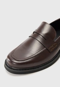 Braune Lederloafers mit glatter Oberfläche, ausgestattet mit einem vorderen Riemen und detaillierter Naht, auf einer schwarzen Gummisohle.