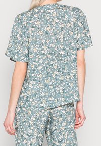 Top a fiori in teal smorzato con maniche corte. Presenta piccoli fiori bianchi e gialli, una vestibilità comoda e un collo rotondo.