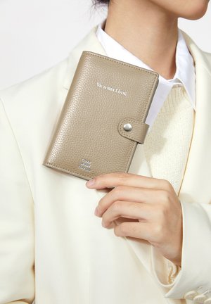 Person in weißer Jacke hält eine geschlossene taupefarbene Lederbrieftasche mit Druckknopf und silbernem Text „Victoria Hyde“ und „Liebe, was du tust“.