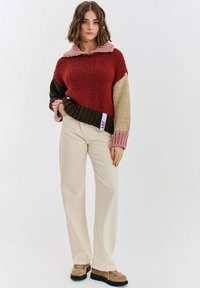 Dziany sweter z blokami kolorów w odcieniach czerwieni, brązu, różu i kremu. Posiada szeroki kołnierz i ściągacze na rękawach. W zestawie z jasnokremowymi spodniami.
