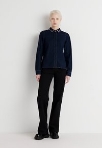 Dark denim button-up shirt with a notched collar featuring silver studs, paired with black flared trousers and chunky black shoes.