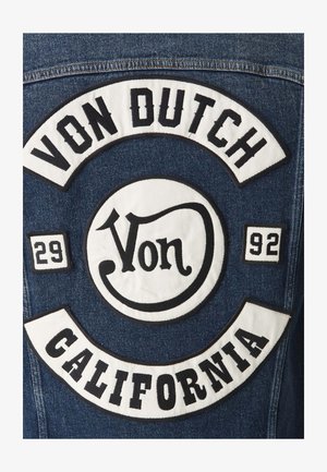 Dos d'une veste en jean avec de grandes patchs blancs et noirs portant les inscriptions « Von Dutch », « California » et les chiffres « 29 » et « 92 ».