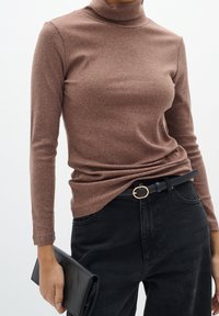 Femme portant un col roulé côtelé marron et un jean noir avec une ceinture noire, tenant une pochette noire dans une main.