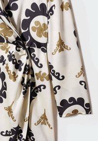 Tissu blanc présentant des motifs noirs et dorés complexes ; comprend des formes tourbillonnantes et des accents décoratifs, avec un détail de taille nouée.