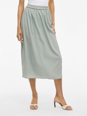 Jupe midi vert clair en tissu fluide avec une taille élastique. Associée à des sandales à talons beiges. Texture lisse et design simple.