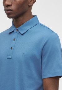 Mann trägt ein hellblaues kurzärmeliges Poloshirt mit drei Knöpfen und kleinem "MK"-Logo auf der Brust.