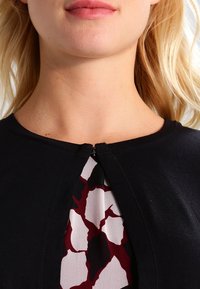 Black top featuring a keyhole neckline with a zipper. Underneath, a patterned fabric in white and burgundy displays abstract shapes.