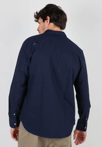 Chemise marine à manches longues avec un col boutonné, dotée d'une patch logo sur l'épaule et d'accents en bouton au niveau du poignet. Texture lisse.