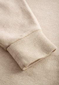 Beige stickad tyg med ribbat manschett. Mjuk textur, något stretchigt, med en jämn yta och ett subtilt melerat mönster genomgående.