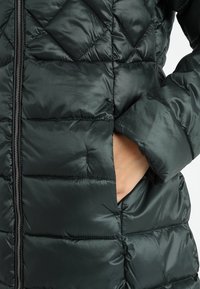 Abrigo acolchado de color verde oscuro con un diseño de rombos, que presenta un cierre frontal con cremallera y bolsillos laterales. La tela tiene una textura brillante y suave.