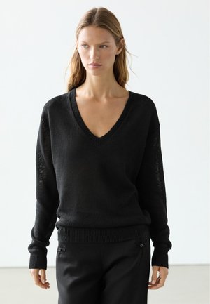 Junge Frau mit langen Haaren, die einen schwarzen V-Ausschnitt-Pullover und schwarze Hose trägt, steht vor einem schlichten hellen Hintergrund.