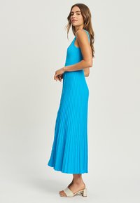 A long, pleated dress in bright blue, featuring a fitted bodice and button details along the front, paired with light-colored heeled sandals.