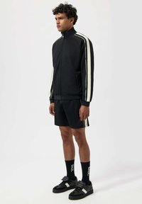 Svart idrottsjacka med vita ränder, kombinerad med svarta shorts. Har dragkedja framtill, strukturerad tyg och matchande svarta sneakers.