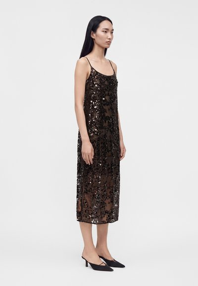 Femme portant une robe midi noire sans manches à sequins et des talons noirs pointus, debout de profil sur un fond clair uni.