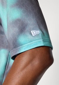 Kortærmet skjorte med en gradient af teal- og gråfarver, med en subtil mønstret tekstur og et hvidt logo på ærmet.