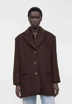 Kobieta modelka z krótkimi kręconymi włosami, ubrana w oversize ciemnobrązowy płaszcz na guziki i jasnoniebieskie dżinsy, stojąca na tle jednolitego jasnego tła.