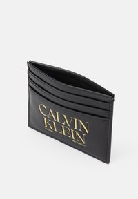 Černý kožený držák na karty s embosovaným zlatým logem "CALVIN KLEIN". Obsahuje více slotů na karty a má elegantní, minimalistický design.