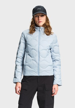 Frau trägt eine hellblaue gesteppte Jacke mit Frontreißverschluss, schwarze Cargohosen und eine schwarze Kappe mit dem "AIGLE"-Logo.