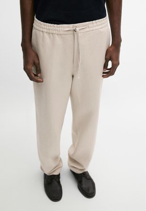 Homme portant un pantalon beige à cordon de serrage et des chaussures noires, debout, les mains détendues le long du corps sur un fond uni.