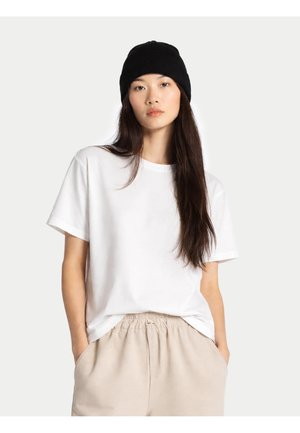 Frau mit langen dunklen Haaren, die eine schwarze Beanie, ein weißes T-Shirt und beige Hose trägt, steht mit den Händen in den Taschen vor einem schlichten Hintergrund.