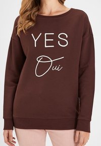 Žena oblečená v hnědém svetru s nápisem "YES Oui" bílou barvou na přední straně, spárovaná se světle zbarvenými kalhotami.