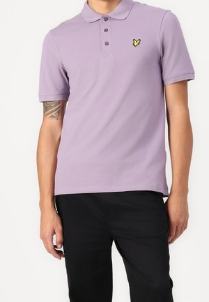Jasnofioletowa koszulka polo z kołnierzykiem, trzema guzikami i żółtym haftowanym logo na lewej piersi. Krótkie rękawy i gładka tekstura.