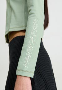 Gaiši zaļš pieguļošs sporta žakets ar ribotām piedurknēm; kreisajā piedurknē ir balta Nike zīmola apzīmējums; gluda tekstūra un īsais dizains.