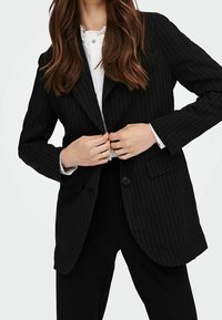 Svart pinstripe blazer med notchade slag, enknapps stängning och två framfickor. Buren över en vit skjorta.
