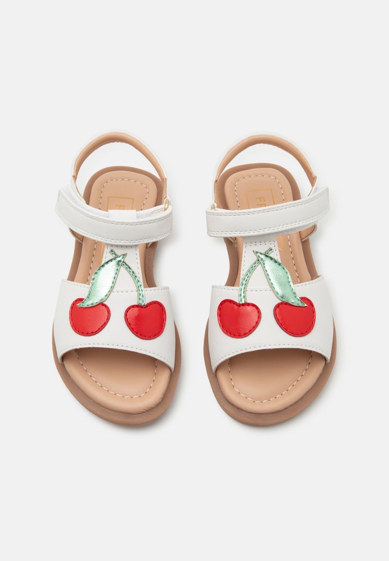 Sandales blanches pour enfants avec des brides à la cheville et des motifs de cerises rouges avec des feuilles vertes sur les brides avant, vues de dessus sur un fond uni.
