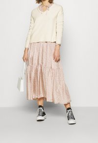 Ecru dzianinowy sweter z dekoltem w serek i wiązaniem; różowa kwiecista, warstwowa długa spódnica; czarne i białe trampki; mała biała torebka.