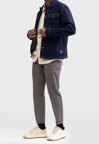 Man bär en marinblå denimjacka över en krämfärgad stickad tröja, rutiga smala byxor, svarta strumpor och beiga sneakers med gummisula.