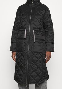 Abrigo largo negro acolchado con cuello alto, cremallera frontal, cintura elástica y dos bolsillos. Tejido con textura y silueta moderna.