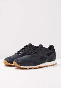Ett par svarta texturerade Reebok-sneakers med gummisula, svarta snören och vita mellansulor, visade på en vit yta.