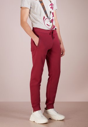 Homme portant un pantalon de jogging bordeaux avec des poches latérales, un t-shirt gris à motif graphique, et des baskets blanches épaisses, debout devant un fond uni.
